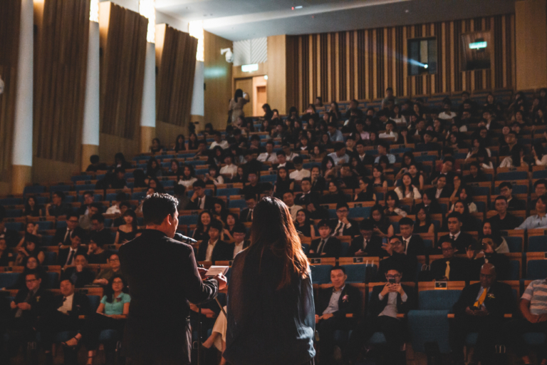 Back view of a man giving a speech to a full lecture hall with a woman standing slightly to his right and behind him, looking at him.
